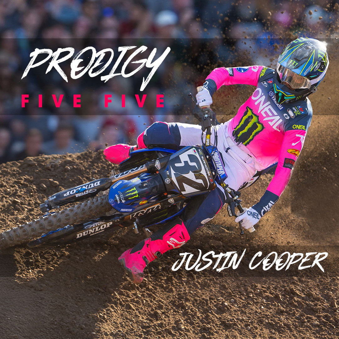Ein Motocross-Fahrer in leuchtend pink-blauer Ausrüstung fährt mit seinem Dirtbike Nummer 32 um eine Kurve. Der Text lautet „PRODIGY FIVE FIVE“ und „JUSTIN COOPER“. Die Szene ist voller Action und Staub.