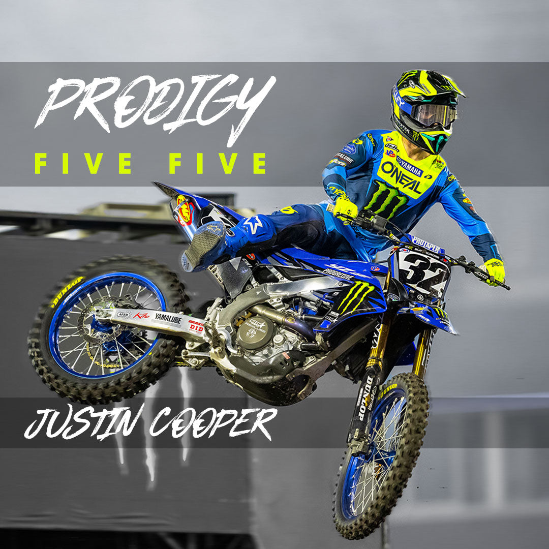 Ein Motocross-Fahrer in blau-gelber Ausrüstung springt auf einem blauen Dirtbike. Der Text lautet „PRODIGY FIVE FIVE“ und „JUSTIN COOPER“. Die Startnummer 32 ist auf dem Bike sichtbar.