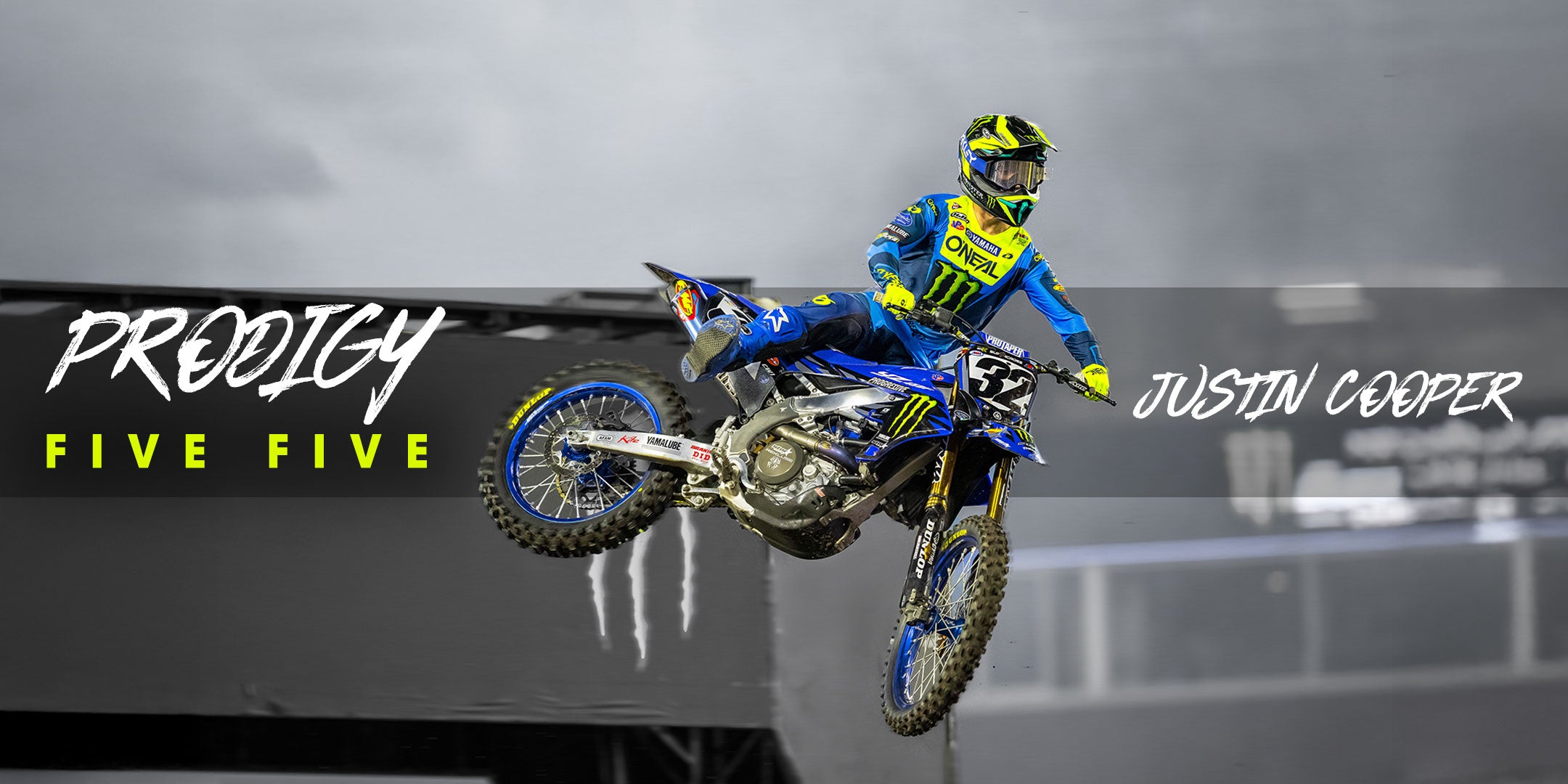 Ein Motocross-Fahrer in blau-gelber Ausrüstung springt auf einem Dirtbike. Der Text auf dem Bild lautet „PRODIGY FIVE FIVE“ und „JUSTIN COOPER“. Der Hintergrund ist unscharf, wodurch die Bewegung hervorgehoben wird.