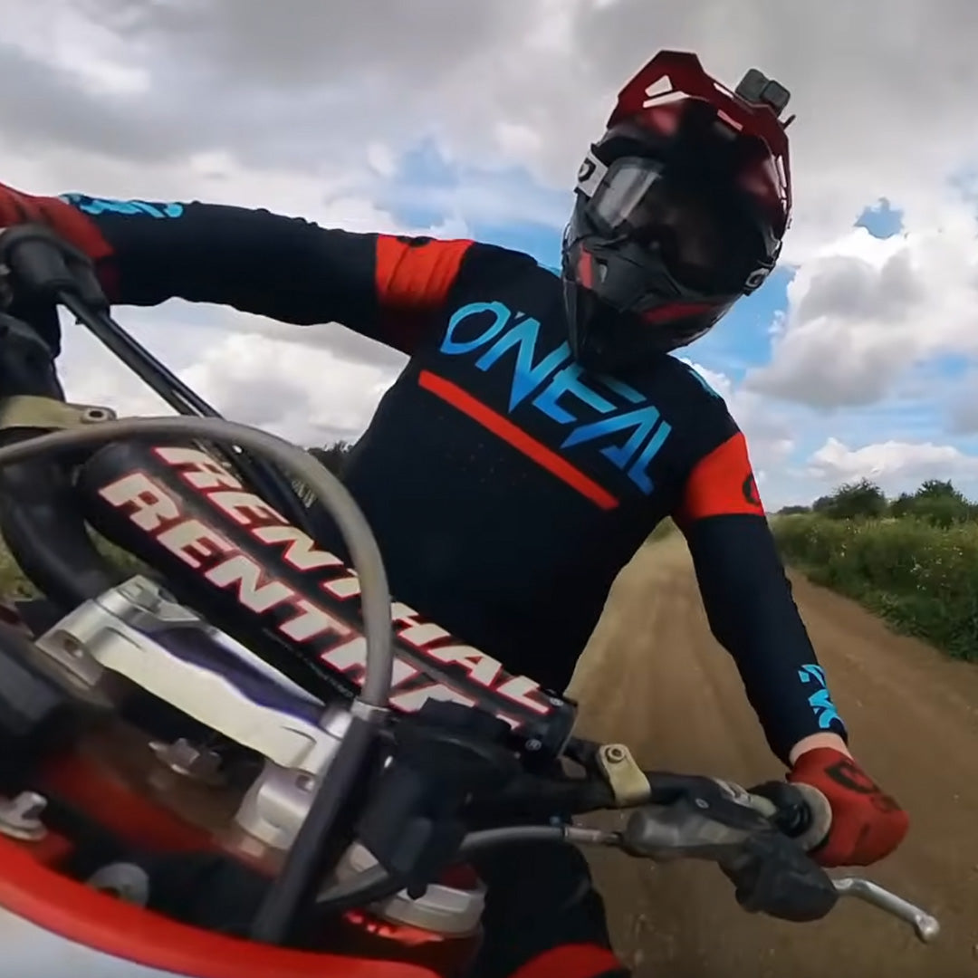 Eine Person mit Helm und O’Neal-Rennausrüstung fährt mit einem Dirtbike auf einer unbefestigten Straße. Das Foto wurde aus einer Untersichtsperspektive mit einem Weitwinkelobjektiv unter einem teilweise bewölkten Himmel aufgenommen.