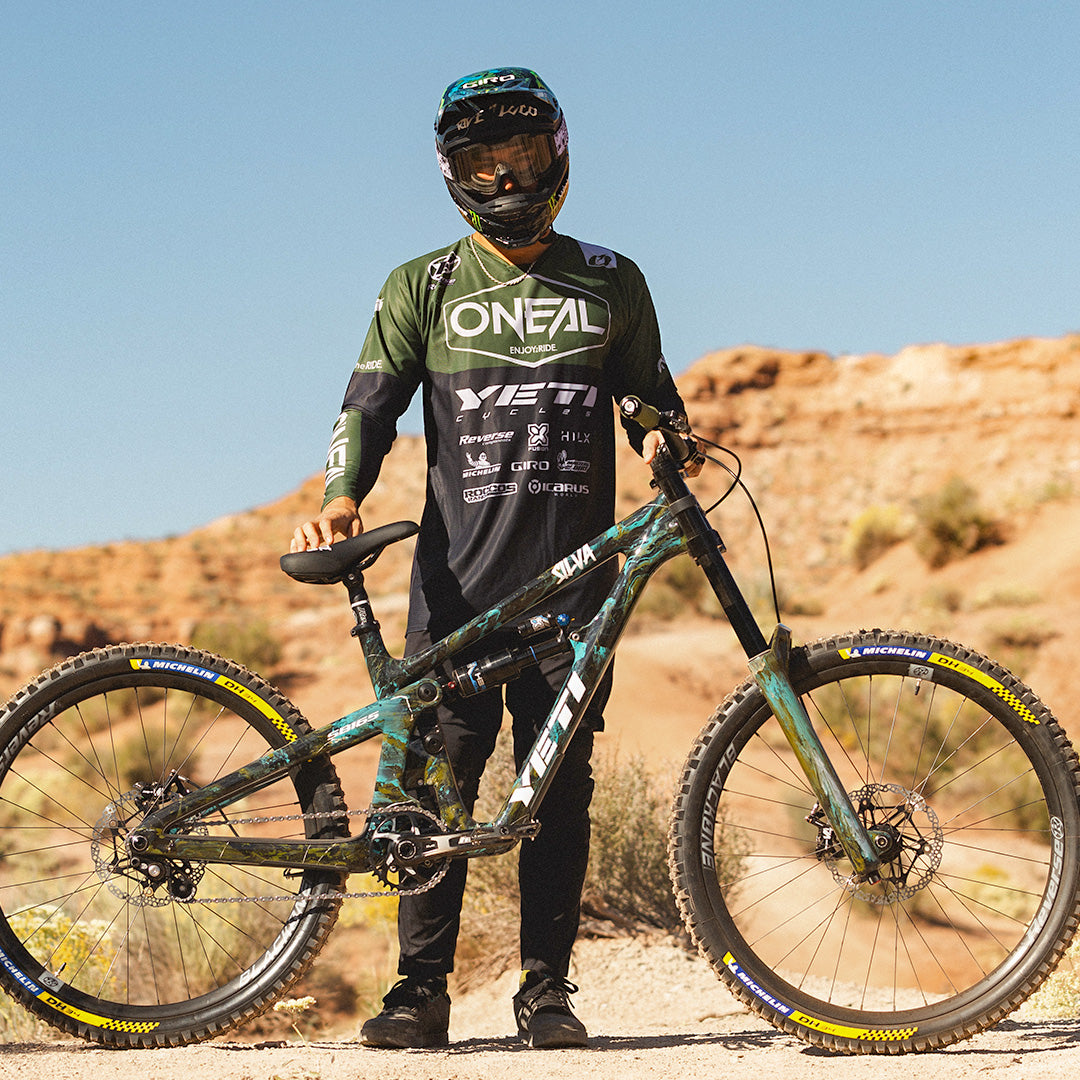 Eine Person in voller Radsportmontur steht im Freien auf felsigem Gelände und hält ein Mountainbike. Sie trägt Helm, Schutzbrille und ein dunkelgrün-schwarzes O'Neal-Trikot. Das Rad hat dicke Reifen und einen robusten Rahmen.