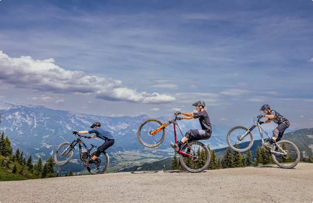Drei Mountainbiker machen Wheelies auf einem Feldweg vor einer Kulisse aus malerischen Bergen und blauem Himmel mit vereinzelten Wolken. Sie sind in Radsportkleidung und Helmen gekleidet und demonstrieren ihre Radfahrkünste vor der üppigen grünen Landschaft, die sich unter ihnen ausbreitet.