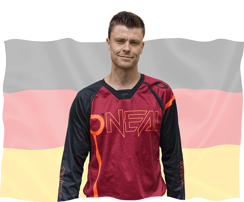 Eine Person in einem rot-schwarzen O'Neal-Shirt steht selbstbewusst vor einer wehenden deutschen Flagge mit horizontalen schwarzen, roten und gelben Streifen. Die Person hat kurze Haare und einen neutralen Gesichtsausdruck.