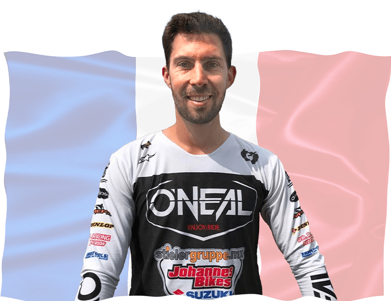 Ein lächelnder Mann in einem Motocross-Trikot steht vor einer großen französischen Flagge. Sein Trikot ist weiß mit schwarzen Akzenten und Logos verschiedener Sponsoren, darunter O'Neal. Der Hintergrund ist eine wellenförmige blau-weiß-rote Trikolore.
