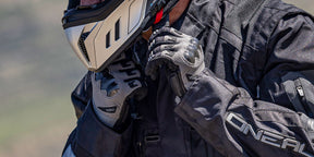 Nahaufnahme einer Person, die eine schwarze Motorradjacke, Reithandschuhe und einen Helm trägt und den Helmriemen befestigt. Der Markenname "O'NEAL" ist auf der Jacke und den Handschuhen zu sehen.