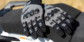 Ein Paar schwarze und graue Motorradhandschuhe mit Knöchelschutz ruhen auf einem schwarzen Sitz. Ein teilweises "READY TO RACE"-Schild ist auf einer weißen Fläche in der Nähe zu sehen.