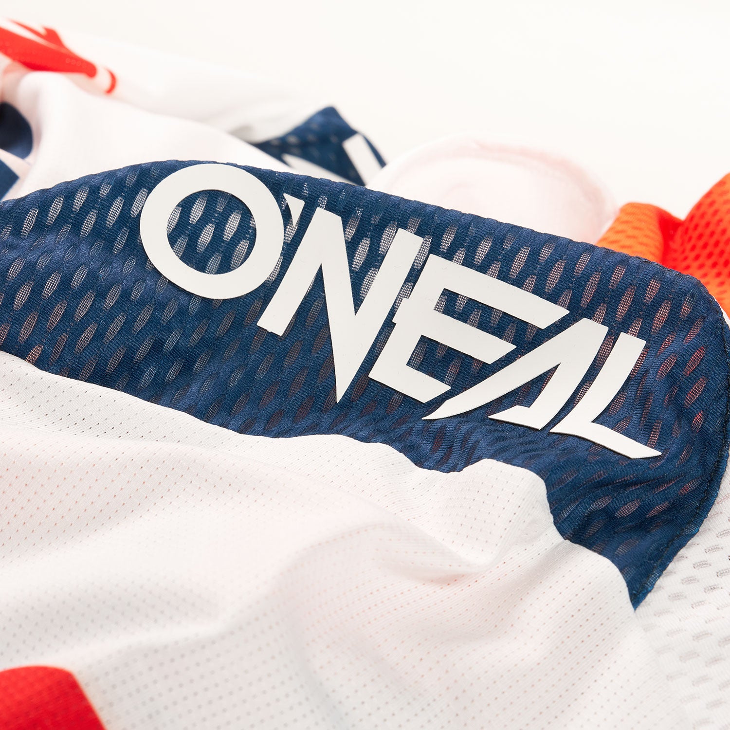 Eine Nahaufnahme eines Sporttrikots mit dem auffälligen weißen „ONEAL“-Logo auf einem dunkelblauen, atmungsaktiven Netzgewebe mit umgebenden Bereichen aus weißem, rotem und orangefarbenem Material.