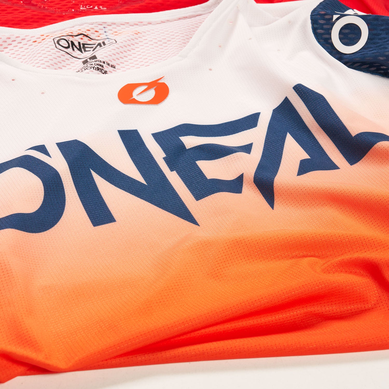 Eine Nahaufnahme eines rot-orange-weißen O'NEAL-Sporttrikots mit dem auffälligen marineblauen Logo der Marke auf der Brust und einem Netzkragen mit sichtbaren Etiketten.