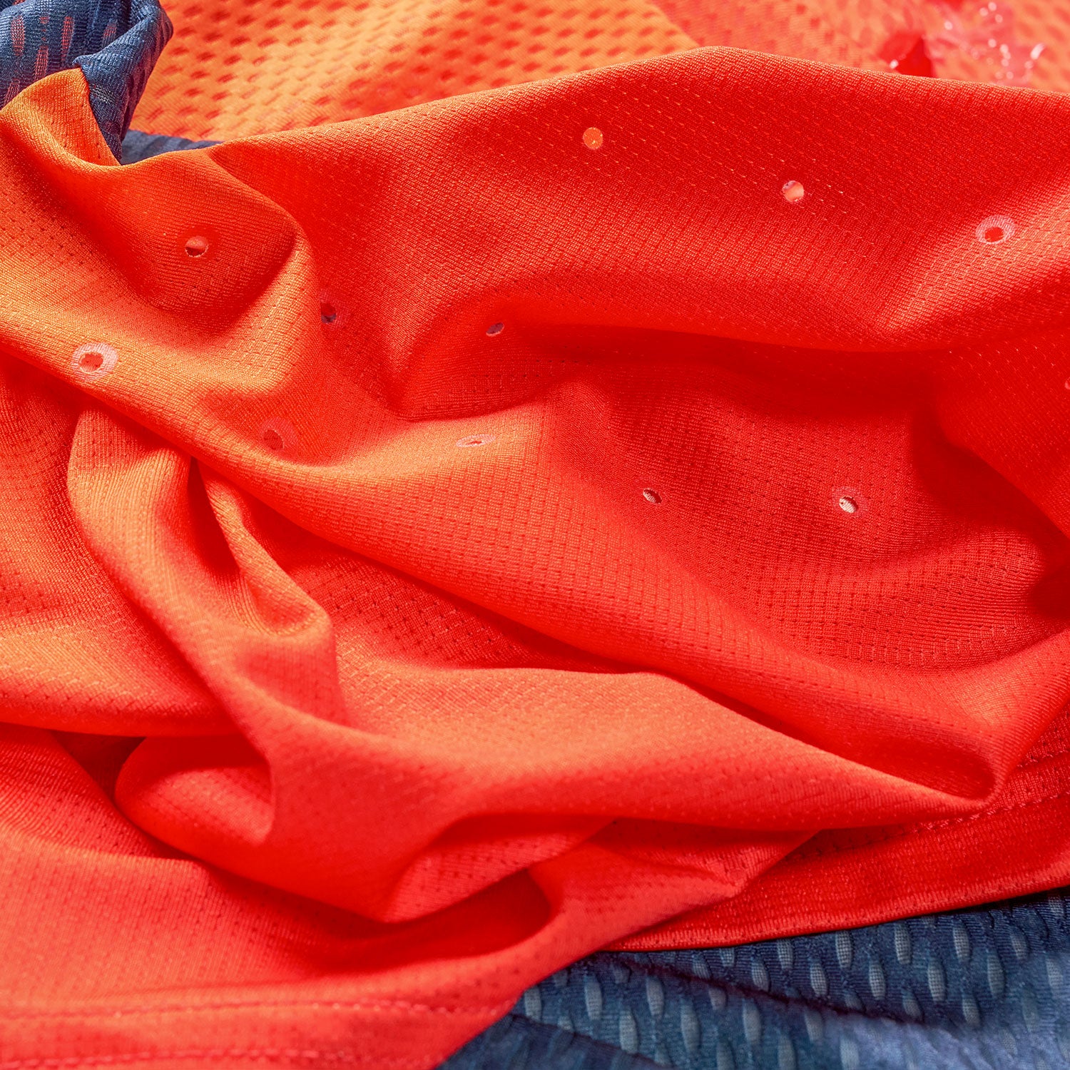 Leuchtend orangefarbener Sportstoff mit kleinen Belüftungslöchern, drapiert und leicht zerknittert, auf einem blau gemusterten Textilhintergrund liegend.