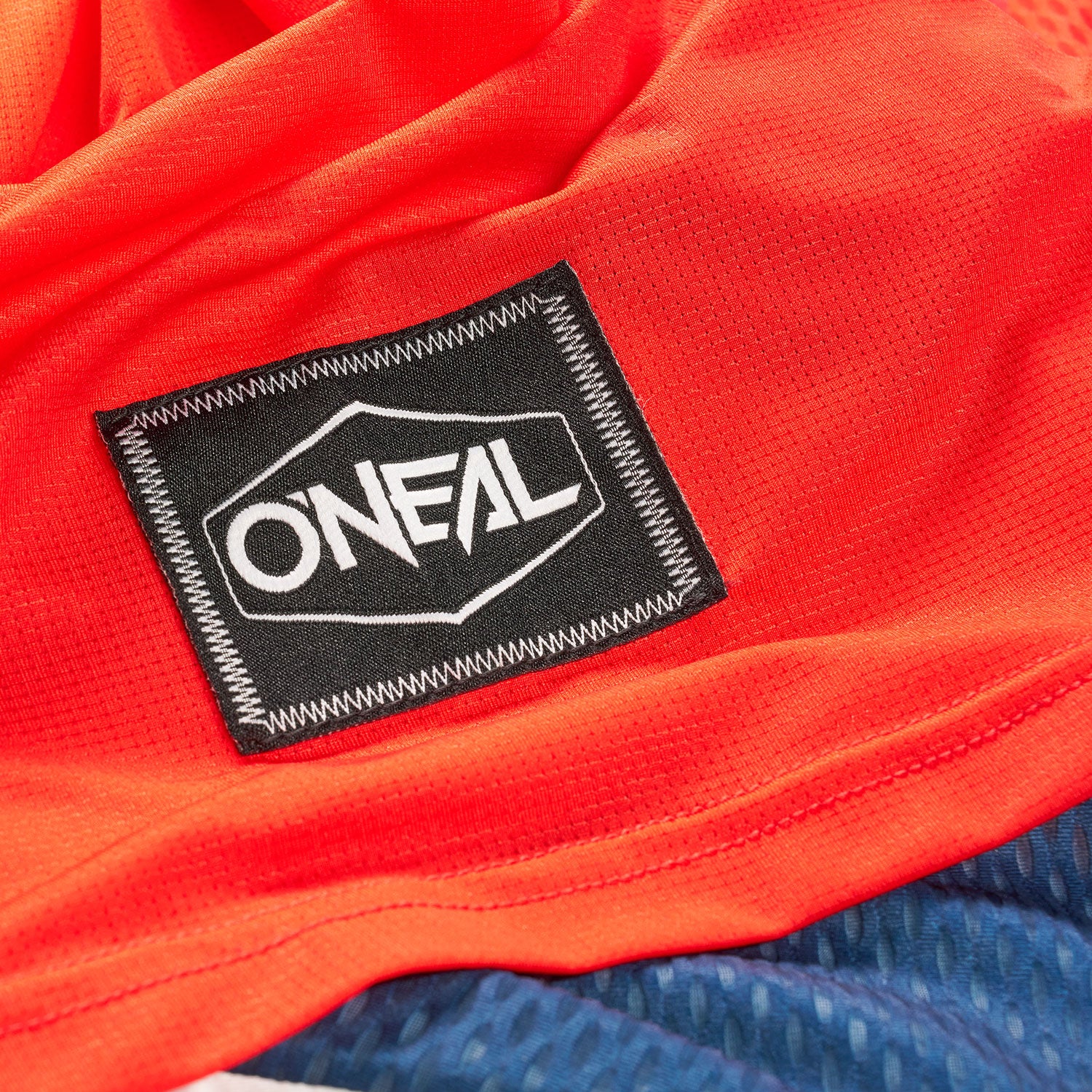 Eine Nahaufnahme eines orangefarbenen Stoffes mit einem aufgenähten schwarzen Patch, auf dem das weiße Logo „ONEAL“ zu sehen ist. Unten im Bild ist teilweise ein blauer Stoff mit einem gepunkteten Muster zu sehen.