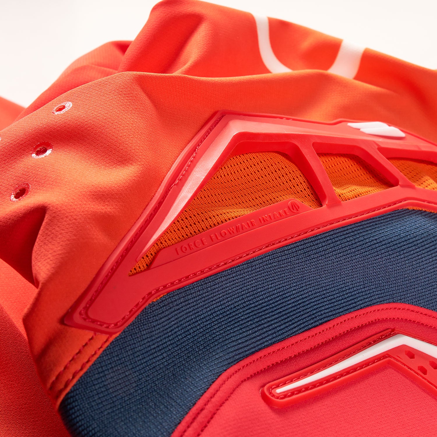 Nahaufnahme eines leuchtend orange-roten Sporttrikots mit einer geometrischen roten Kunststoffstruktur, Netzgewebe und dem sichtbaren Text „FORCE FLOW AIR“. Auch etwas blauer Stoff ist sichtbar.