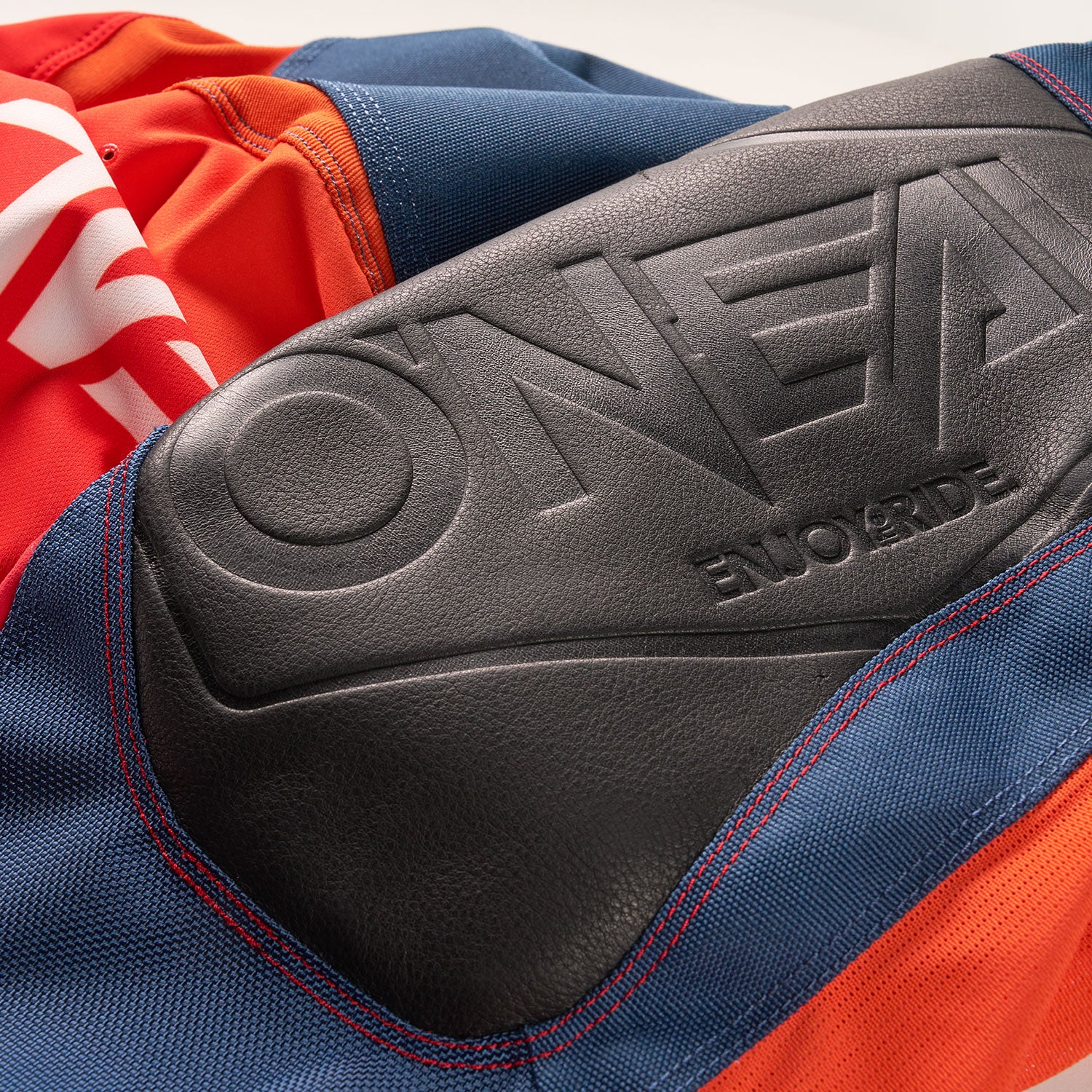 Nahaufnahme eines schwarzen gepolsterten Abschnitts mit der Prägung „O'NEAL ENDURO RIDE“, befestigt an einem Kleidungsstück aus rotem, blauem und orangefarbenem Stoff mit sichtbaren Nähten.
