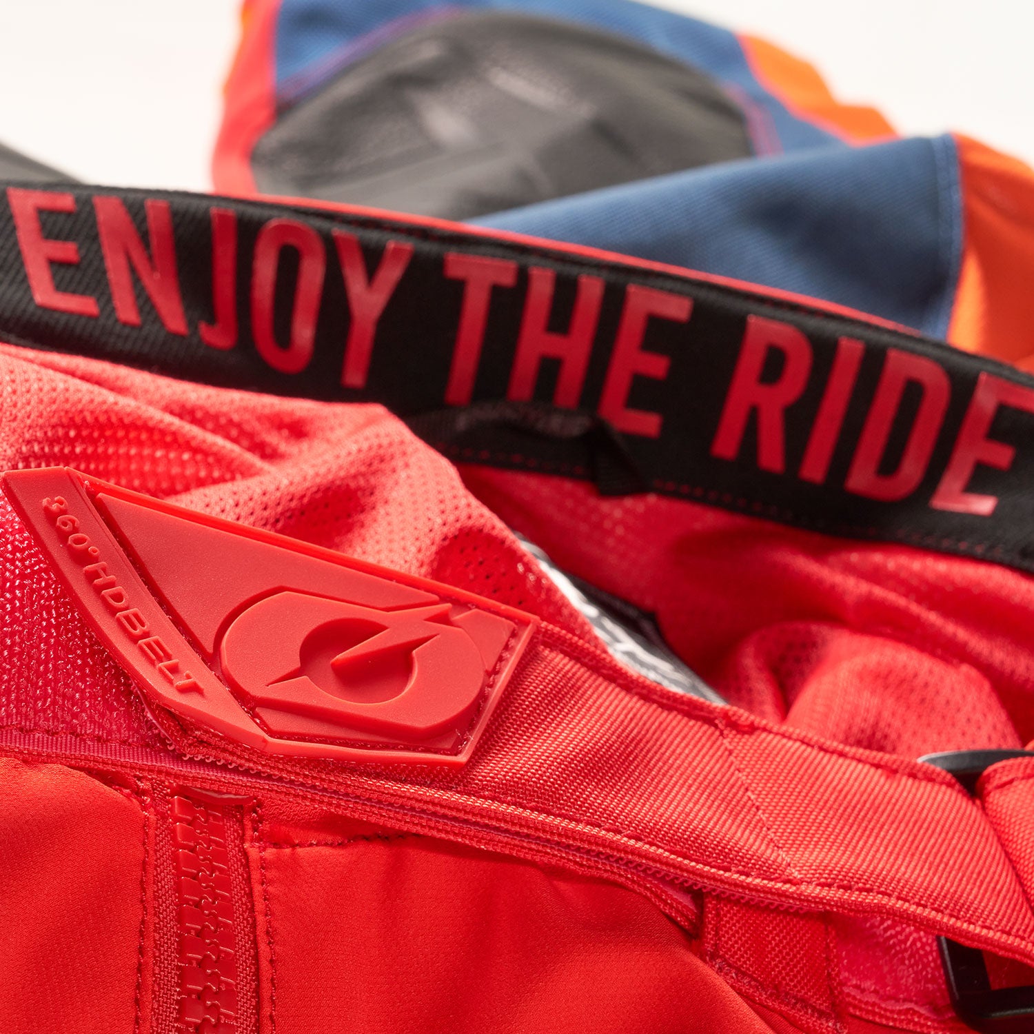 Nahaufnahme farbenfroher Sportbekleidung mit einem roten Hosenbund mit dem Satz „ENJOY THE RIDE“ in fetten Buchstaben und einem roten Logoaufnäher aus Gummi mit dem Markennamen „Troy Lee Designs“ und einem stilisierten Buchstaben T.