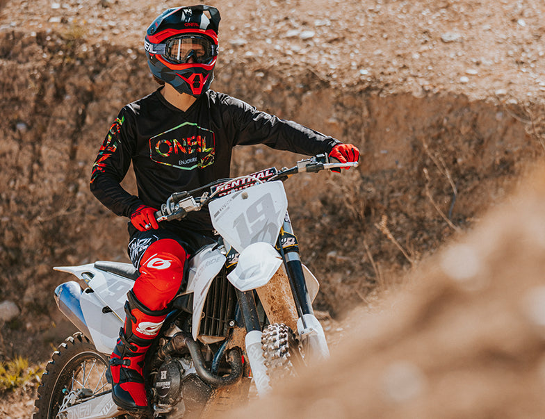 Ein Motocrossfahrer in rot-schwarzer Ausrüstung mit Helm und Schutzbrille sitzt auf einem Dirtbike mit weißem Nummernschild. Im Hintergrund ist eine unbefestigte Strecke mit unebenem Gelände und Felsen zu sehen. Der Fahrer blickt zur Seite und scheint fahrbereit zu sein.